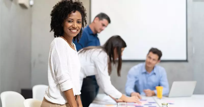 Mulher sorrindo em ambiente de trabalho colaborativo com colegas ao fundo.