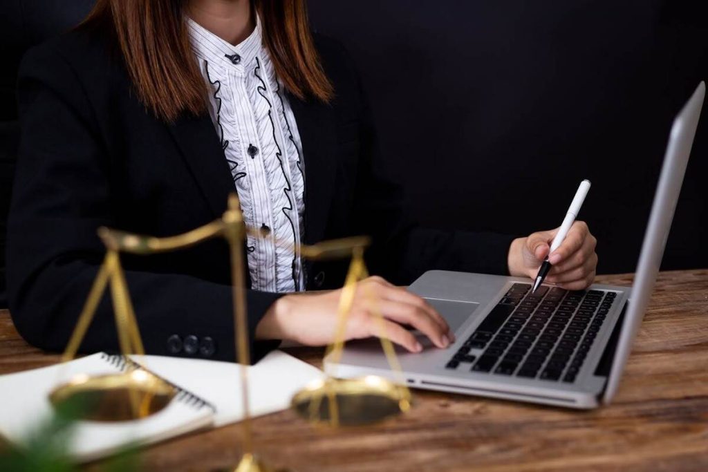 Mulher advogada usando laptop em escritório com balança da justiça ao lado, simbolizando a advocacia moderna e a tecnologia.