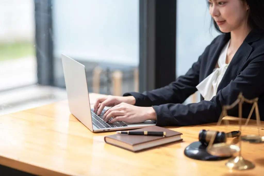 Mulher usando laptop em ambiente de trabalho, com mesa de madeira, bloco de notas e itens de escritório como caneta e martelo de juiz.