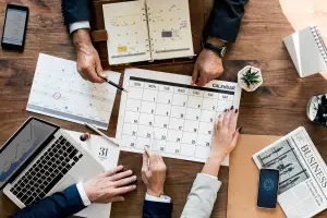 Mesa com um calendário no centro e mãos de alguns trabalhadores de terno sobre ele, com um notebook à esquerda representando software para controle de prazos judiciais.